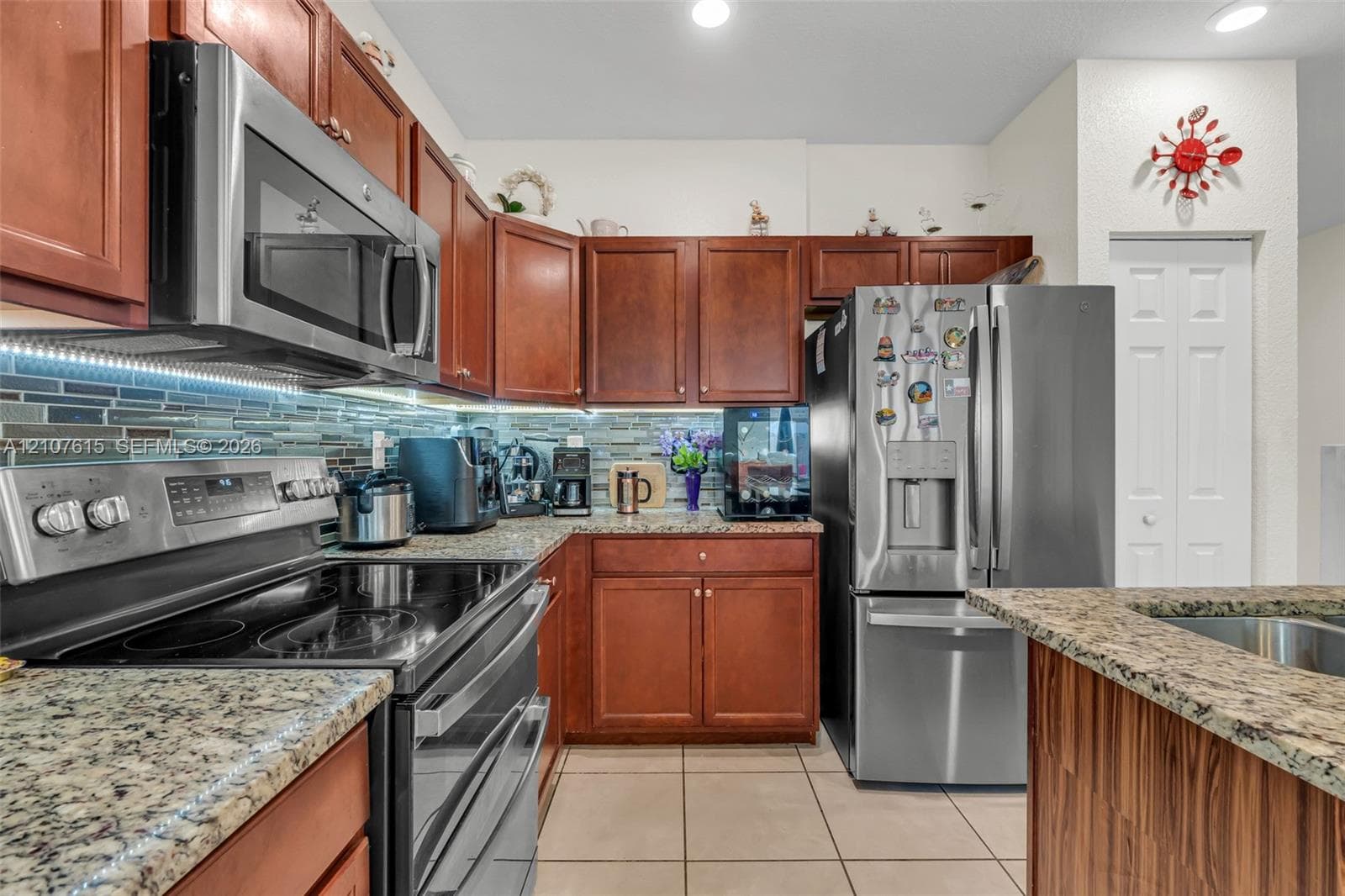Kitchen featuring rich-toned wood cabinetry, stone-finish countertops, stainless steel appliances, multi-tonal glass tile backsplash, and ceramic floor tiling