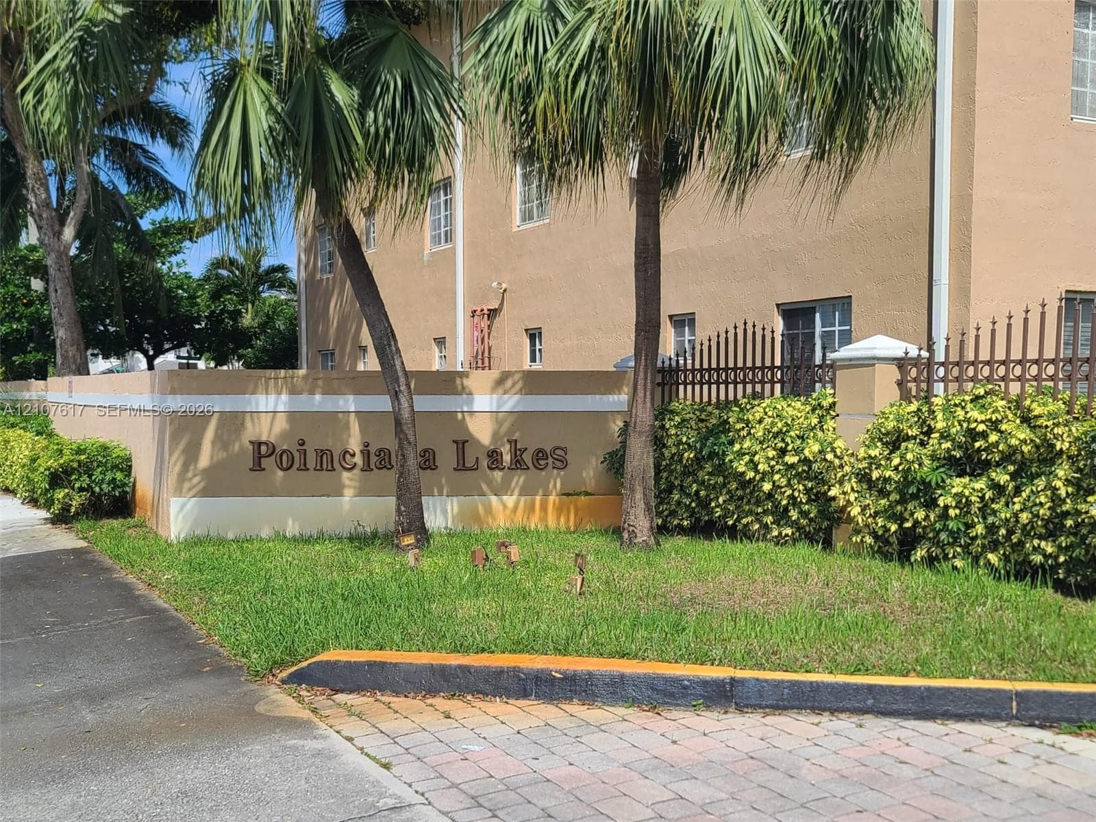 Stucco monument signage featuring ornamental metal fencing, concrete paver drive entry, and mature palm landscaping