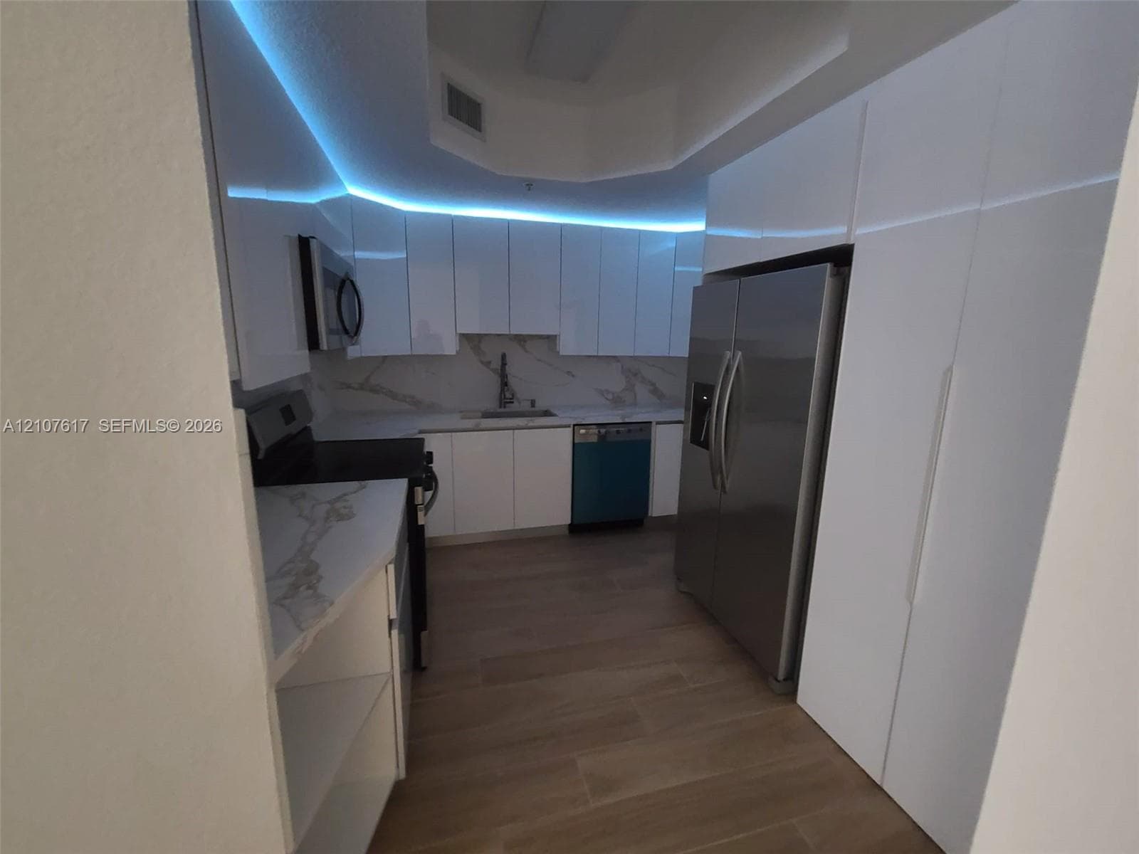 Kitchen featuring high-gloss white cabinetry, stone-patterned countertops, integrated lighting, stainless steel appliances, and wood-finish flooring