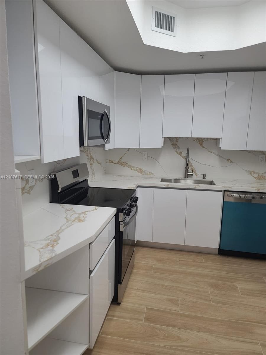 Modern kitchen featuring high-gloss white cabinetry, stone-finish countertops with matching backsplash, stainless steel appliances, wood-finish flooring, and a recessed ceiling skylight
