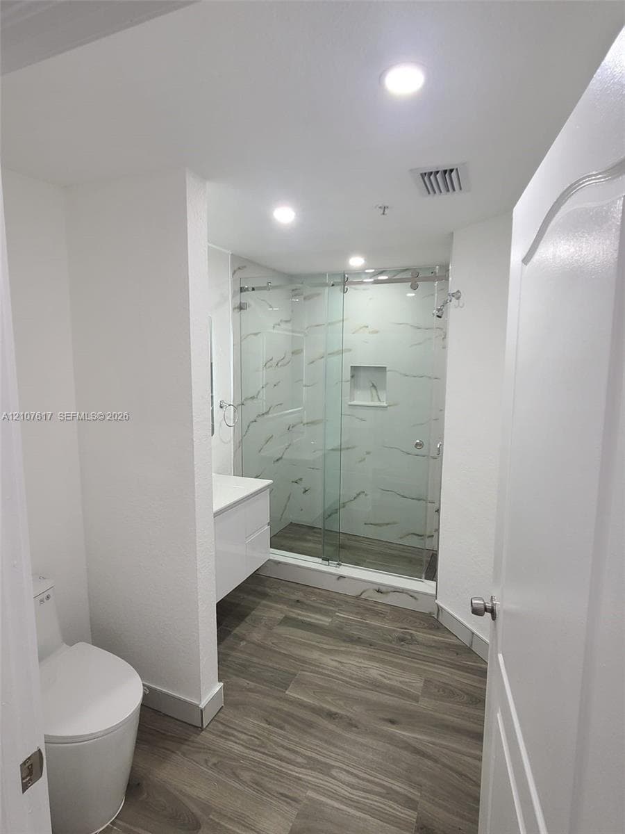 Walk-in glass-enclosed shower with tiled surround and built-in niche, wood-finish flooring, white floating vanity, and recessed lighting