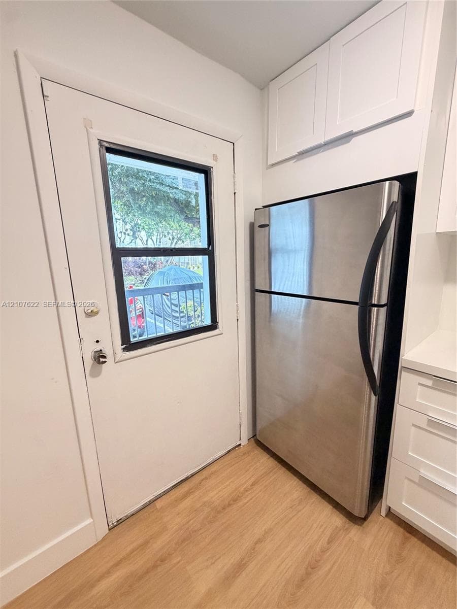 Stainless steel refrigerator, white cabinetry with overhead storage, wood-finish flooring, and a single-light exterior door
