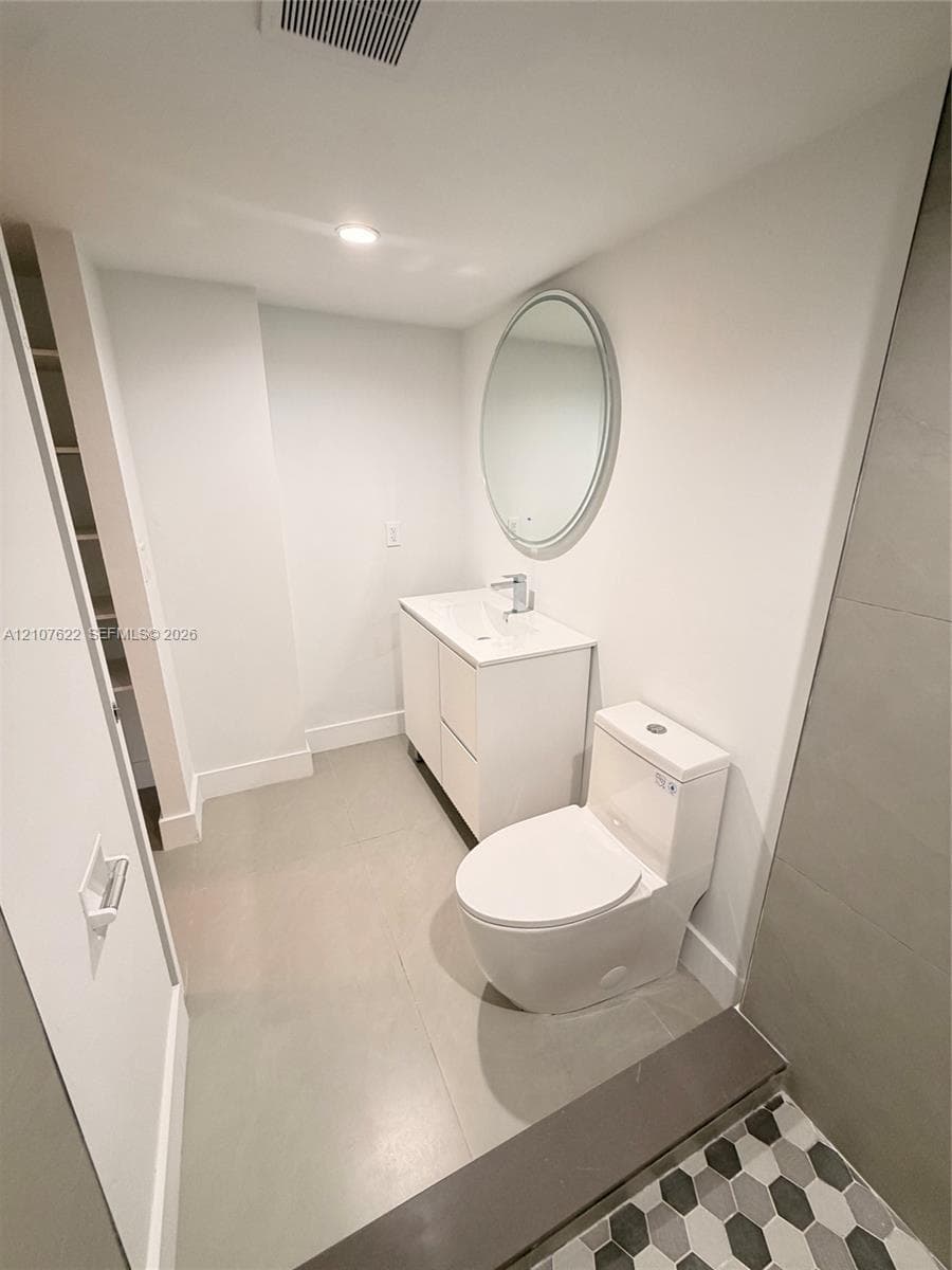 Bathroom featuring a white vanity cabinet, rectangular integrated sink, oval wall-mounted mirror, dual-flush toilet, and hexagonal floor tile
