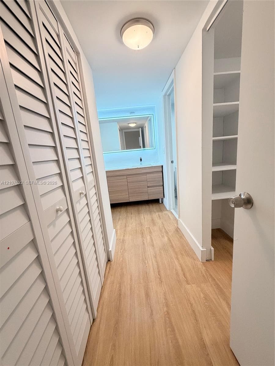 Wood-finish flooring, white louvered-style closet doors, built-in open shelving, vanity with integrated cabinetry, and an illuminated vanity mirror