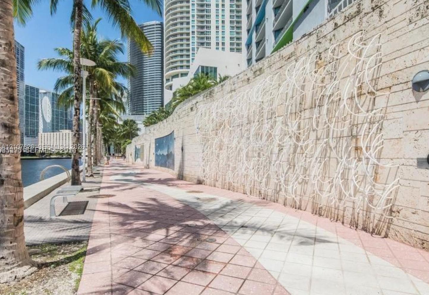 Waterfront promenade featuring a textured stone perimeter wall, decorative wall-mounted metal art, paver stone walkway, and mature palm trees