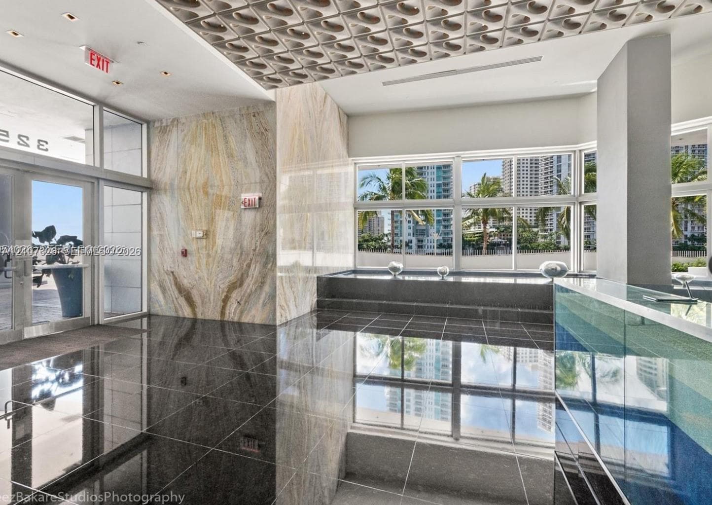 Lobby interior featuring large-format dark stone flooring, book-matched stone wall cladding, a geometric-patterned drop ceiling, floor-to-ceiling glass paneling, and a custom built-in reception desk
