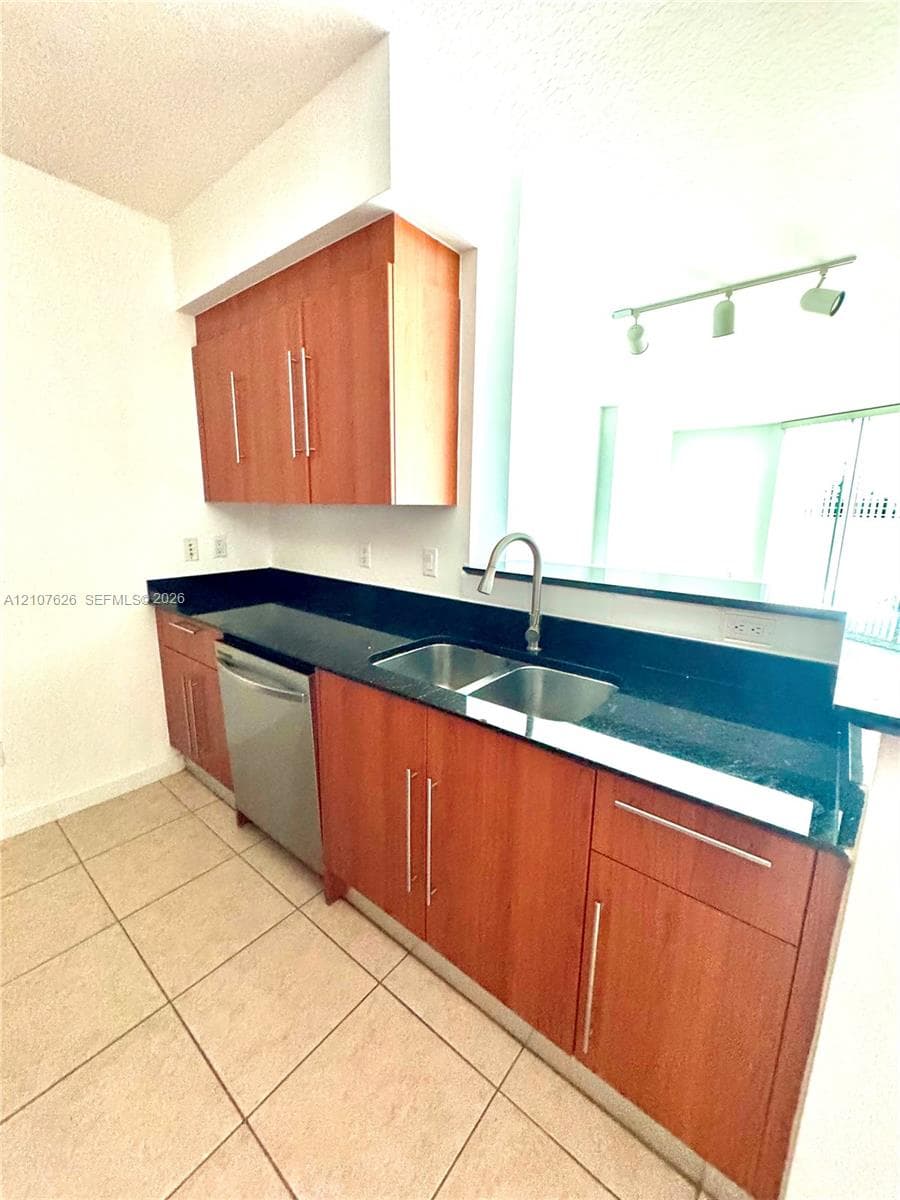 Kitchen featuring wood-finish cabinetry, dark solid-surface countertops, dual-basin stainless steel sink with pull-down faucet, integrated stainless steel dishwasher, and track lighting