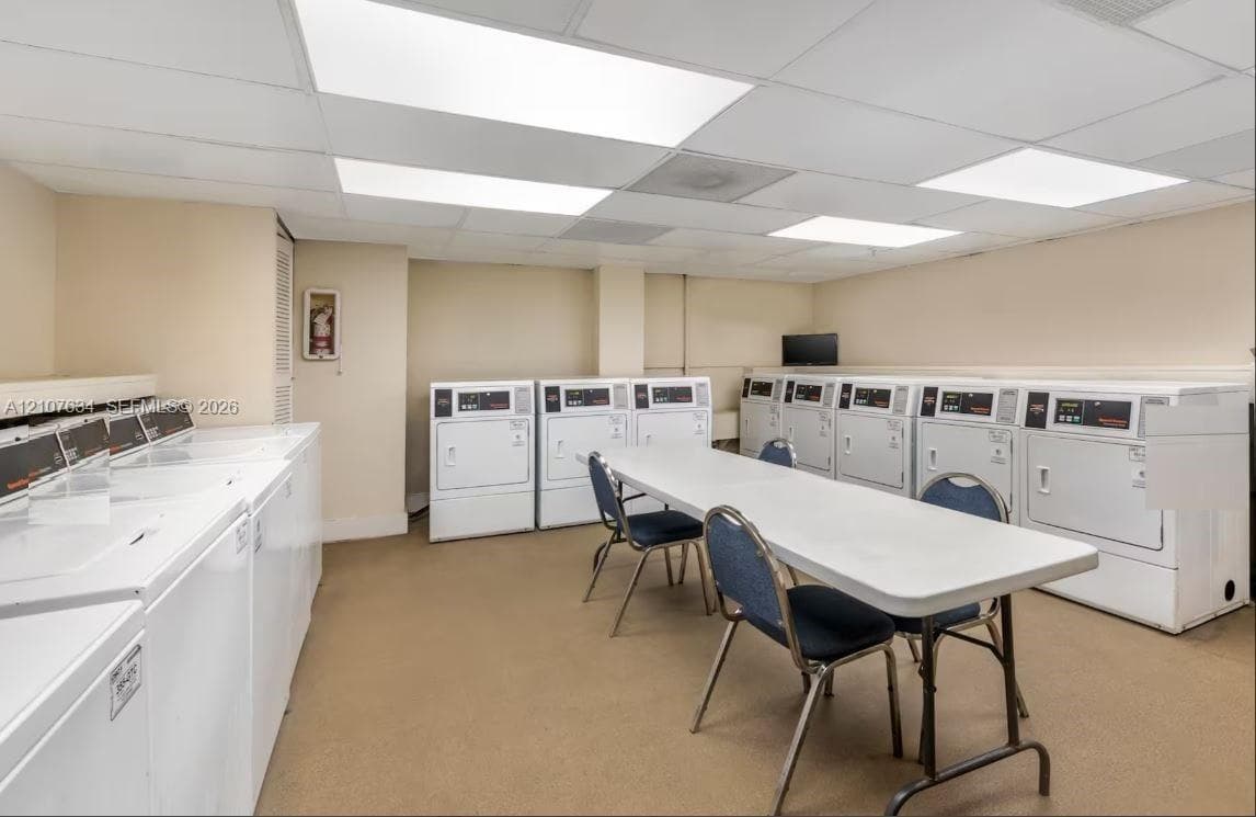 Laundry facility featuring multiple white washing machines, commercial-grade clothes dryers, and recessed panel lighting