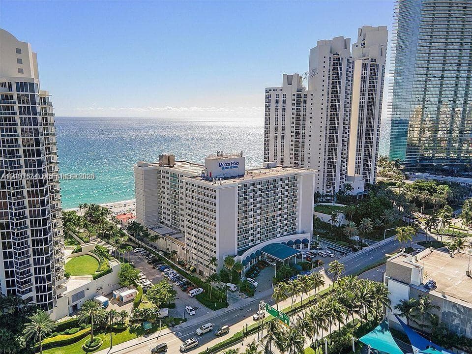 Aerial perspective of a high-rise residential complex featuring oceanfront positioning, direct access to the coastline, landscaped grounds, and proximity to major roadways