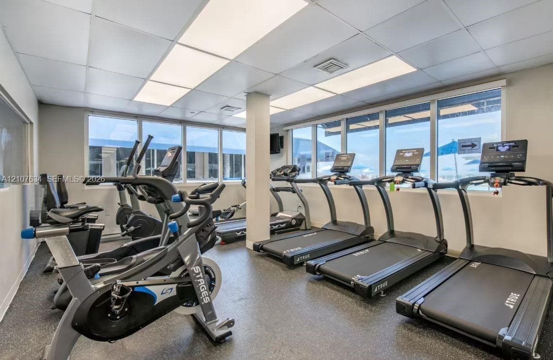 Fitness room featuring rubberized flooring, expansive windows, fluorescent drop ceiling lighting, and a support column