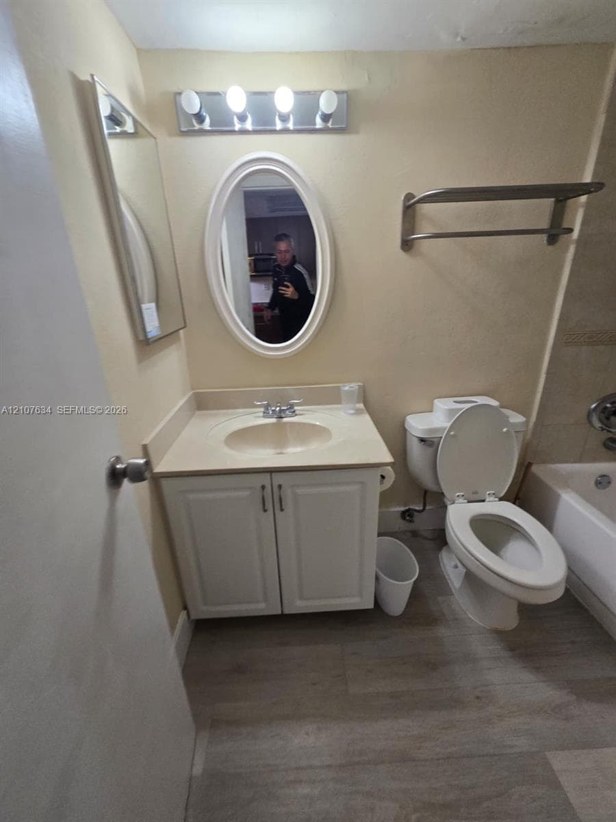 Bathroom vanity with a white cabinet, integrated countertop, and oval-shaped wall mirror, complemented by wood-finish flooring, a wall-mounted towel rack, and four-bulb vanity lighting