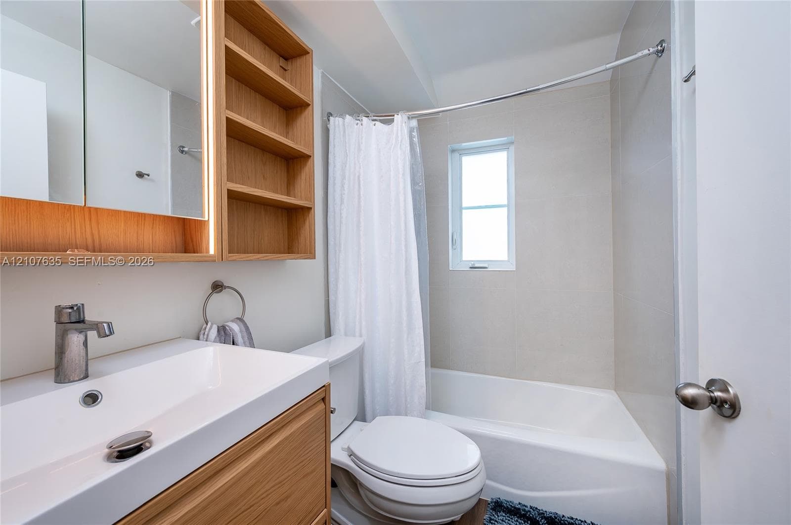Wood-finish vanity with integrated rectangular sink and side shelving, wall-mounted lighted medicine cabinet, and combination tub and shower with light-toned wall tile