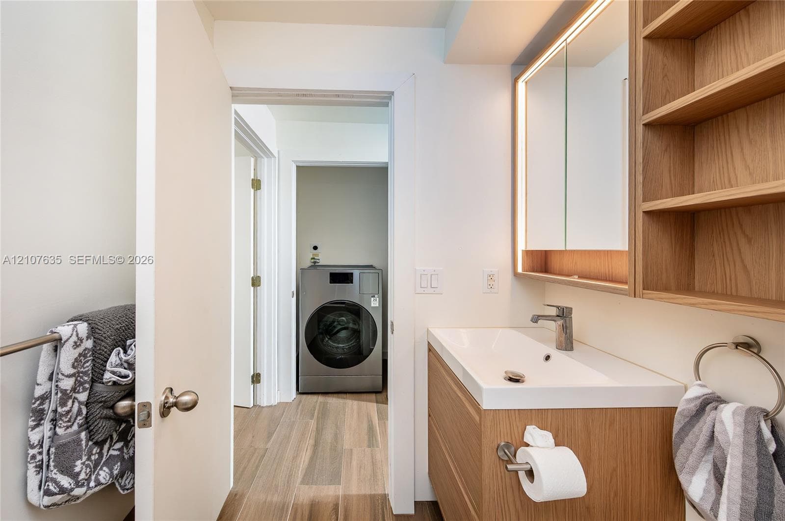 Bathroom featuring a floating vanity with a light wood finish, integrated white basin, illuminated medicine cabinet, built-in open shelving, and stainless steel washer and dryer