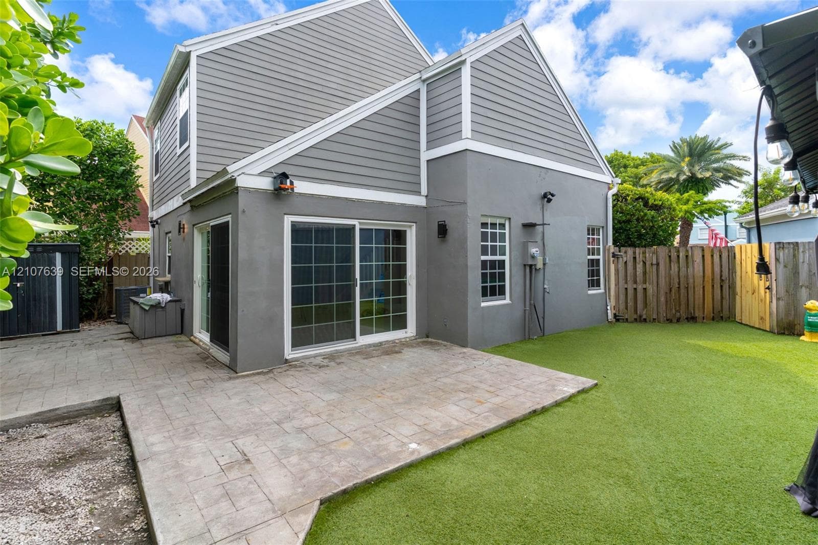 Exterior featuring grey horizontal siding and smooth-finish stucco, paver patio, sliding glass doors, and artificial turf lawn enclosed by wood fencing