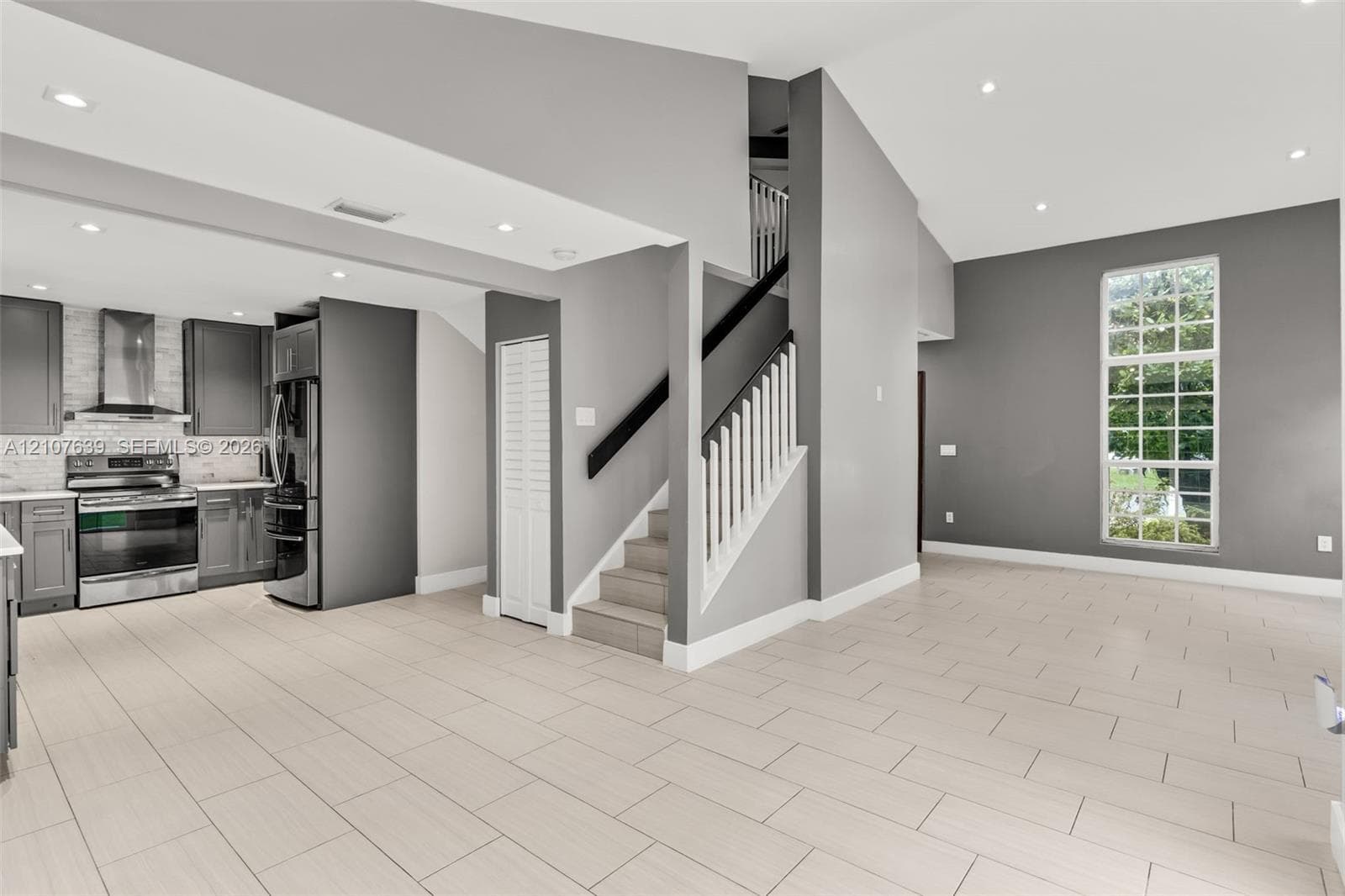 Open-concept interior featuring vaulted ceilings, recessed lighting, tile flooring, a staircase with a white spindle railing, and a kitchen equipped with stainless steel appliances and custom cabinetry