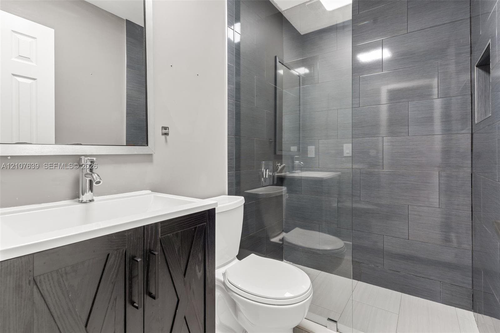Bathroom featuring a vanity with dark wood-finish cabinetry, a glass-enclosed shower with charcoal-toned tile surround, built-in shower niche, and rectangular wall mirror