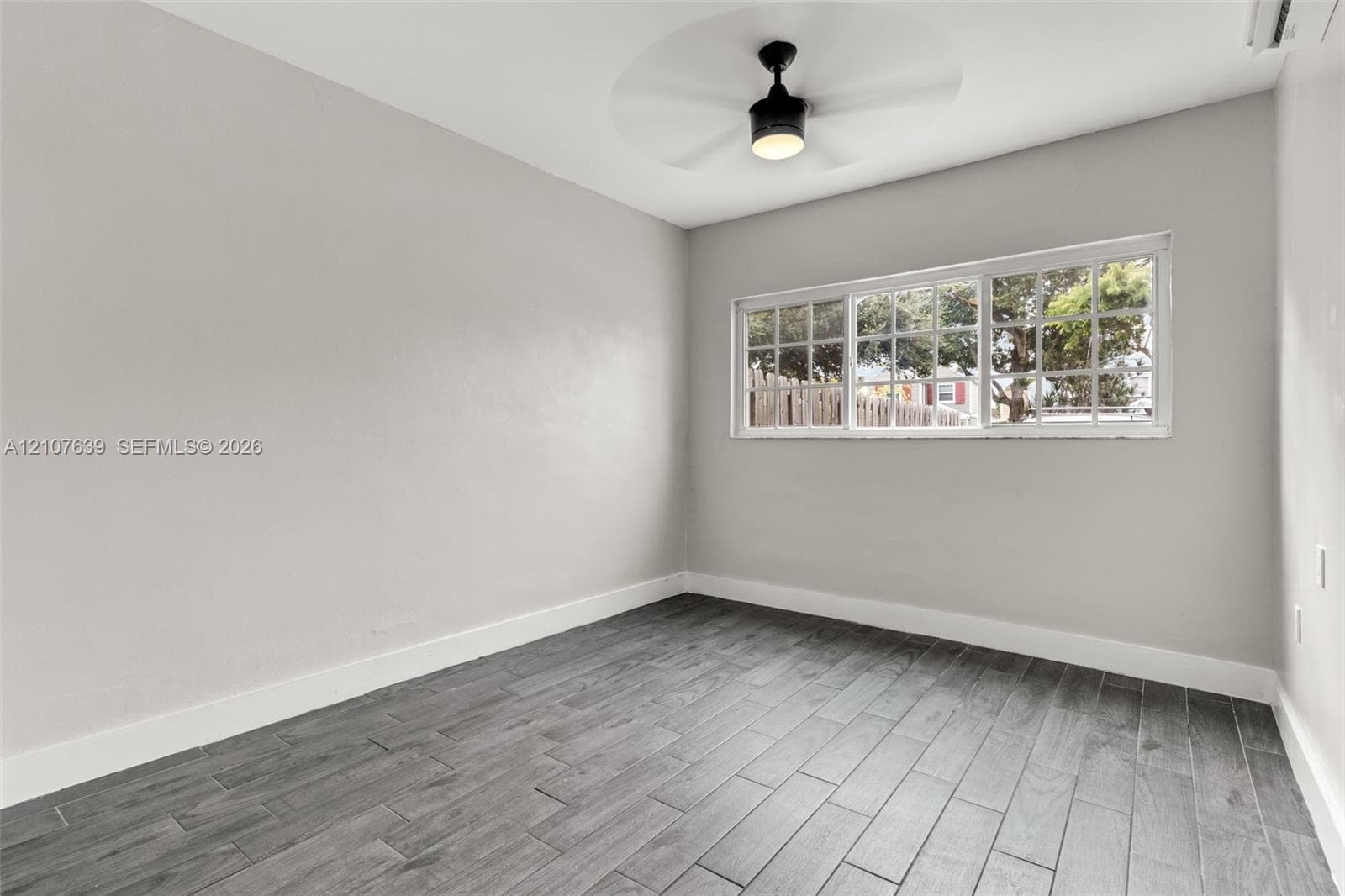 Room interior featuring grey wood-finish flooring, neutral wall paint, a multi-pane horizontal window, and a ceiling-mounted light fixture with a fan