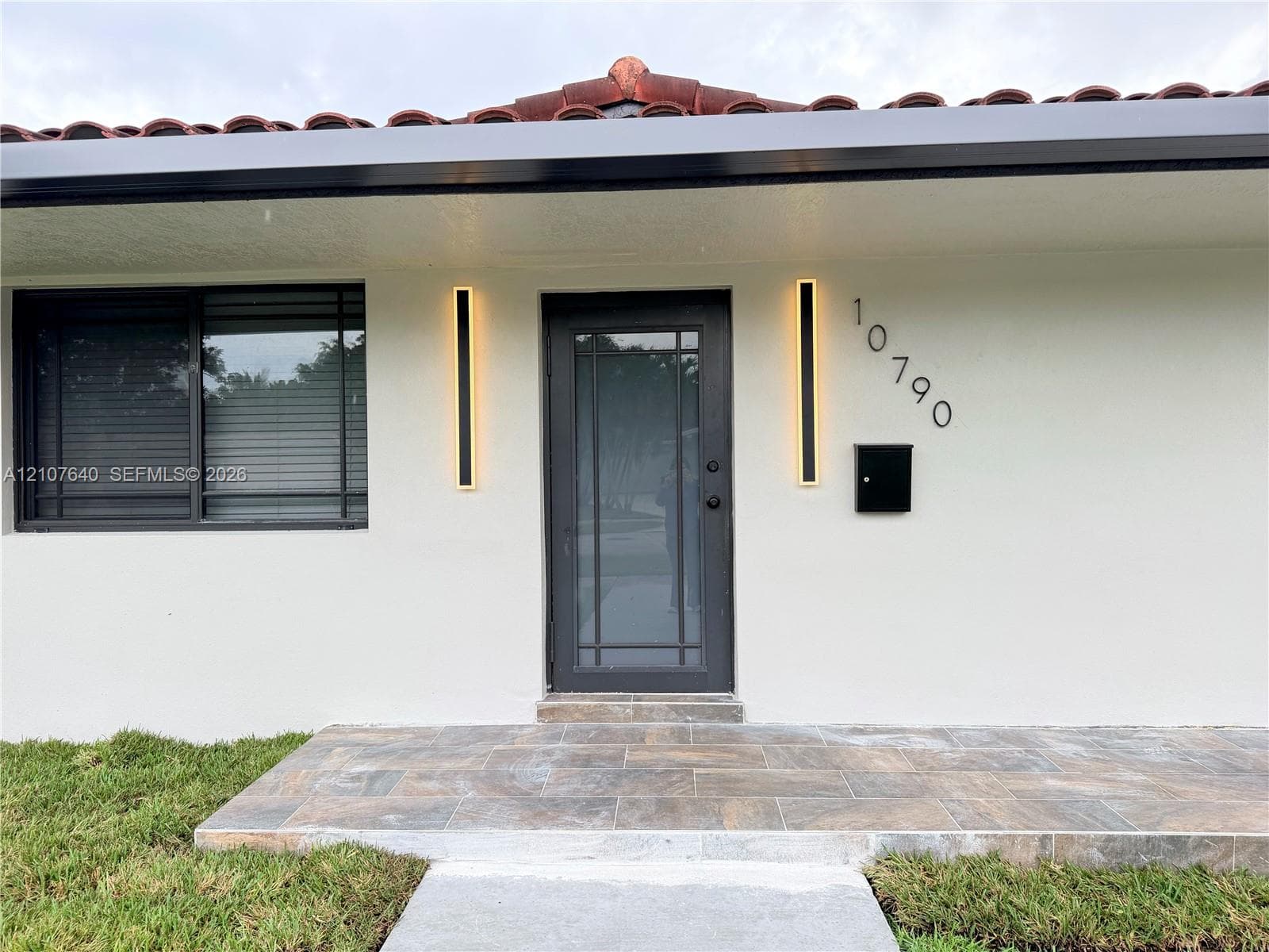 Exterior entryway featuring a terracotta tile roof, stone-finish porch, multi-pane glass-inset door, and vertical linear wall-mounted lighting