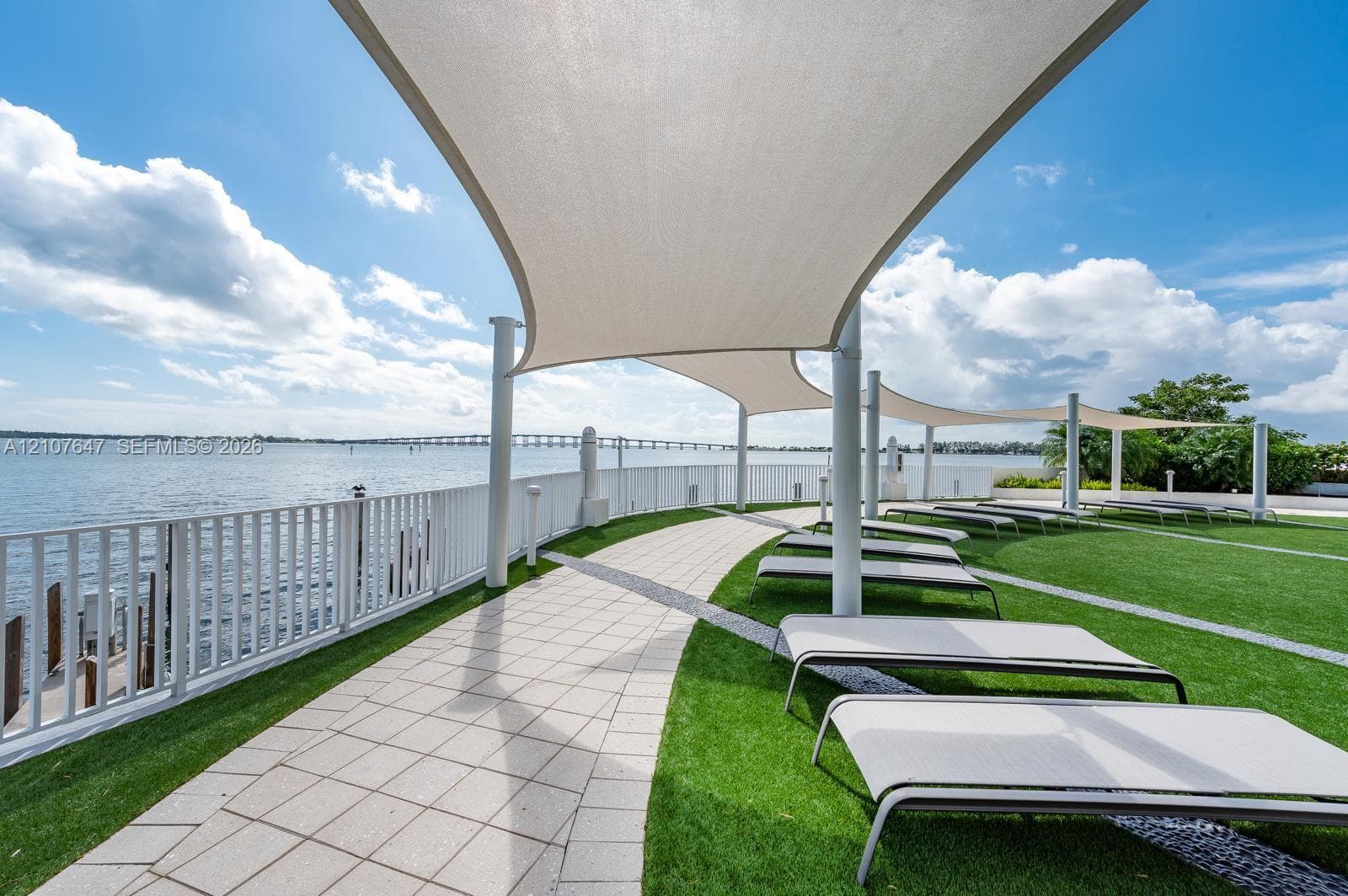 Waterfront patio featuring expansive white sail shade structures, synthetic turf lawn, stone-paver walkway, and white metal railing