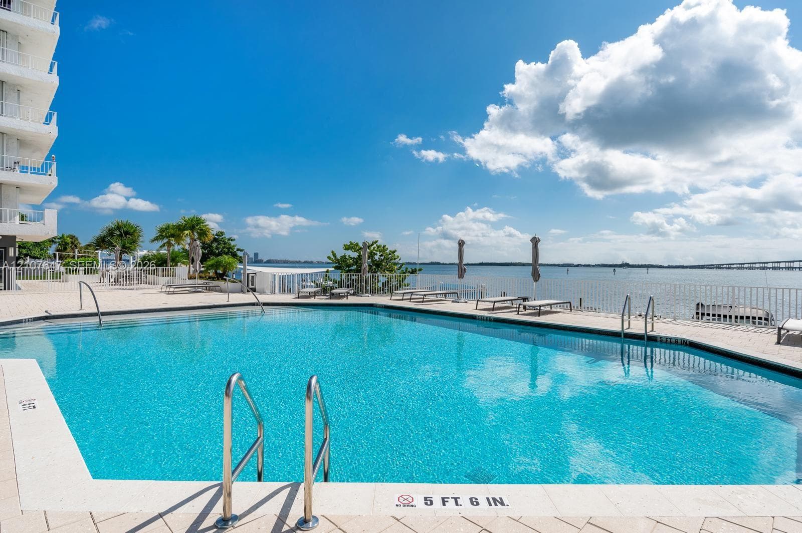 Swimming pool featuring metal handrails, stone paver decking, and direct water frontage with a protective perimeter fence