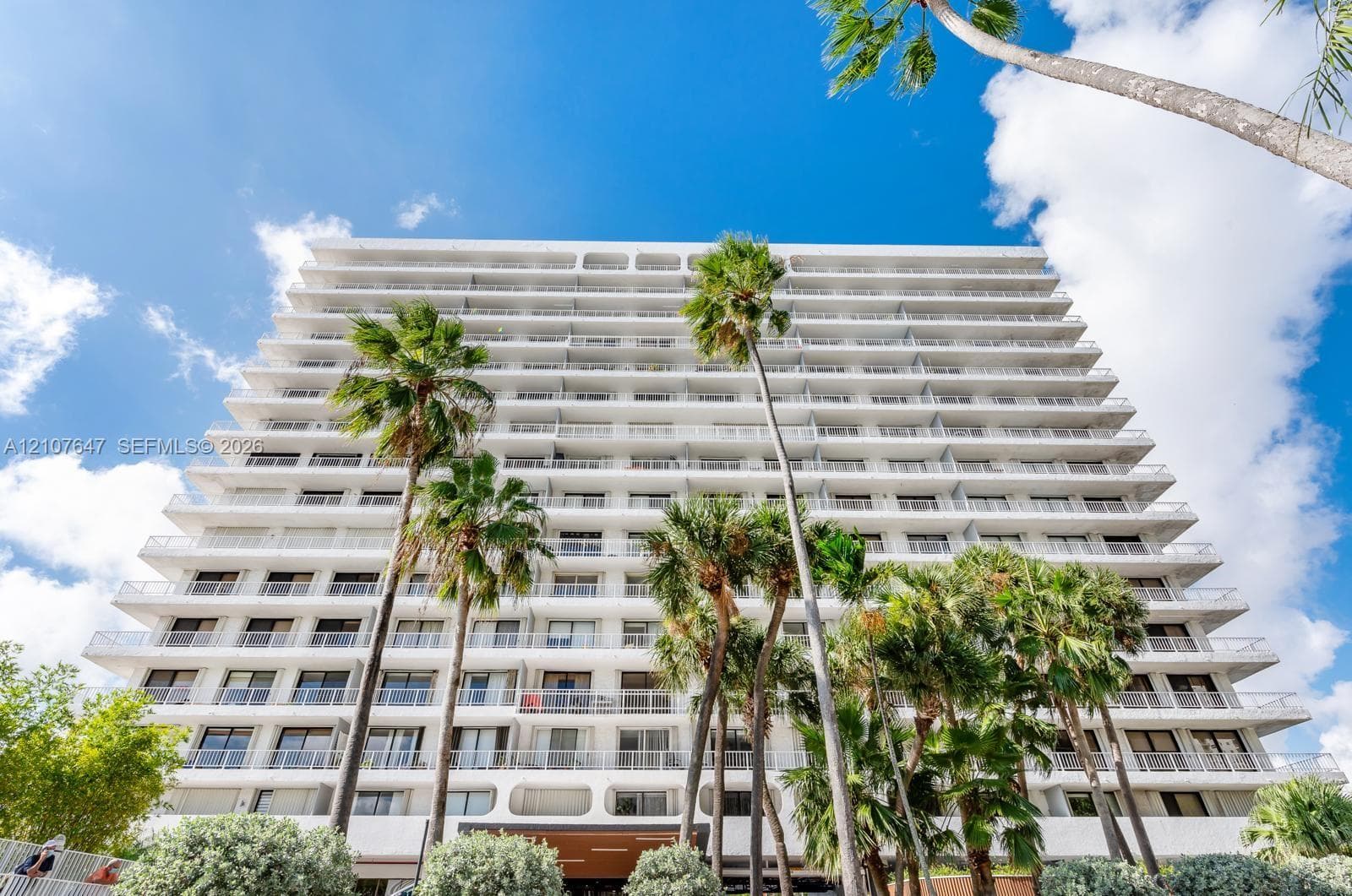Multi-story high-rise building featuring private balconies, floor-to-ceiling glass sliding doors, and structural concrete framing, accented by mature palm trees