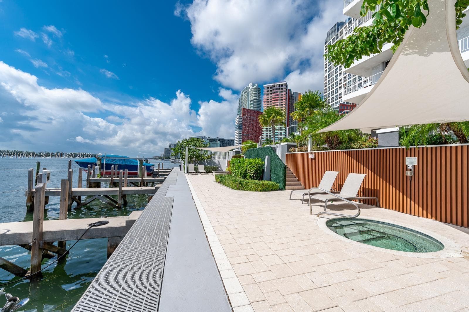 Waterfront terrace featuring stone paving, an in-ground spa, a private boat dock, vertical wood-slat fencing, and a retractable tension canopy