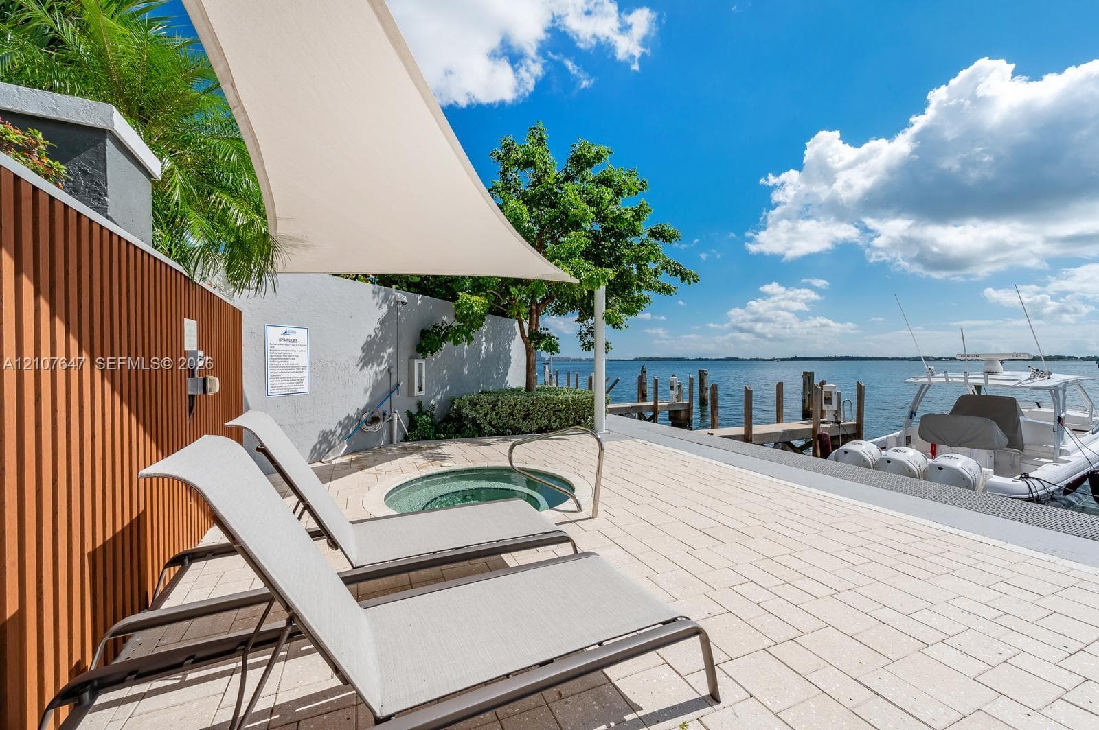 Waterfront terrace featuring stone paver flooring, a built-in spa, structural shade sail, and a private wooden boat dock