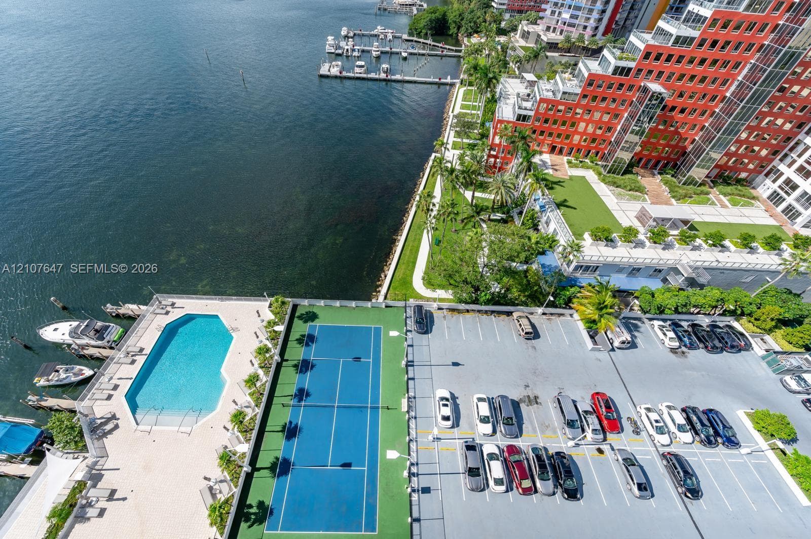 Aerial perspective of property amenities including a waterfront swimming pool, a hard-surface tennis court, and dedicated on-site parking