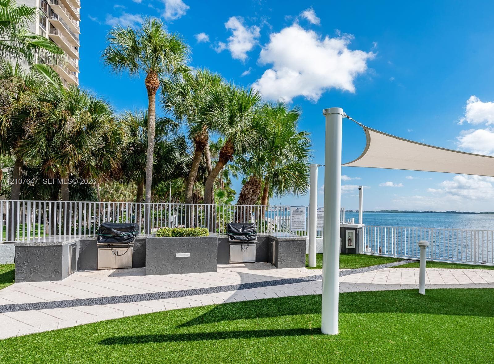 Outdoor cooking area featuring stone-finish counters, built-in stainless steel grills, artificial turf lawn, decorative paver walkway, and a tension shade structure overlooking the water