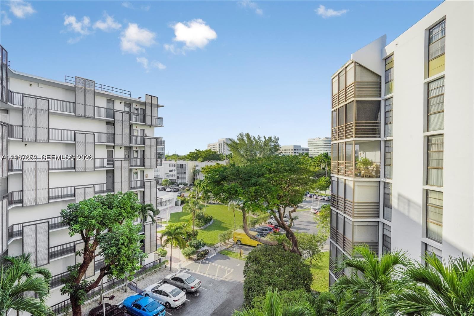 Multi-story residential complex featuring white stucco facades, grid-patterned exterior metal screening, glass-enclosed balconies, and landscaped grounds with mature trees