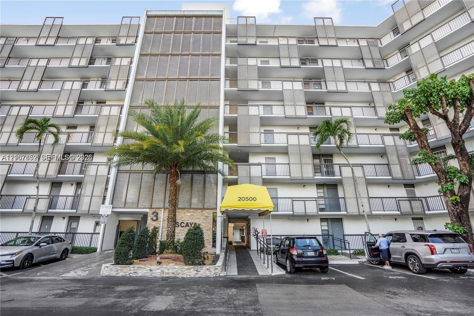 Mid-rise building exterior featuring a stone-clad facade, private balconies with metal railings, decorative metal screening, and an integrated covered entrance