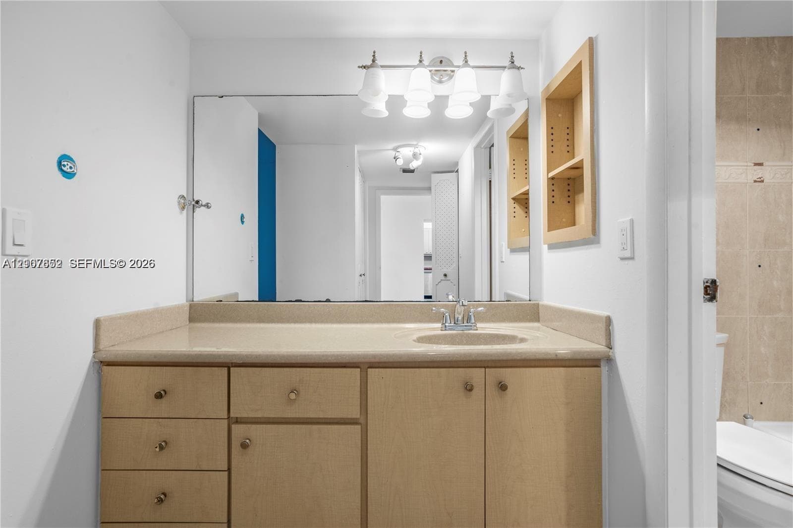 Vanity featuring wood-finish cabinetry, solid-surface countertop, integrated sink, large wall-mounted mirror, and four-light overhead fixture