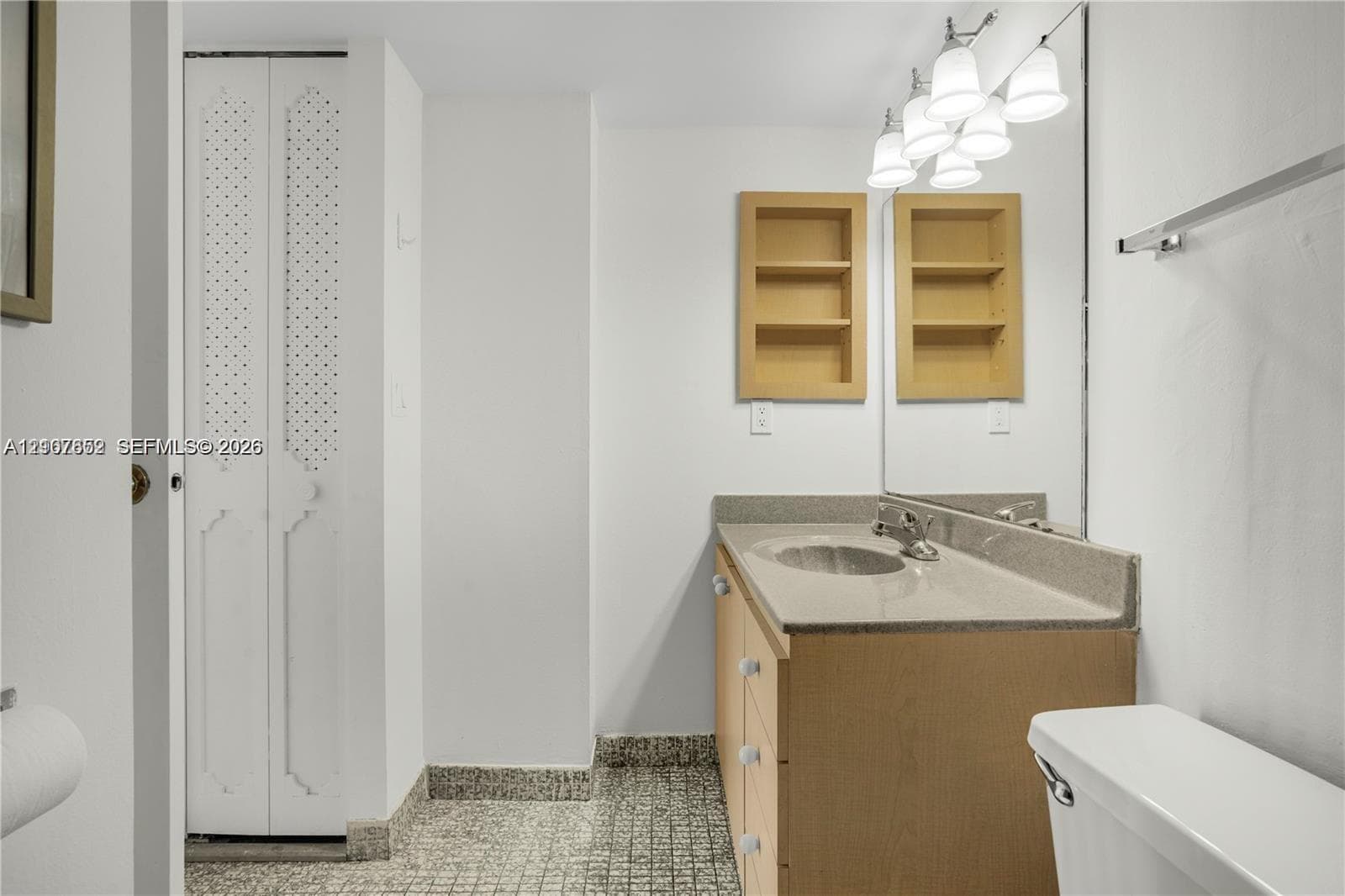Bathroom vanity with a solid-surface countertop and integrated sink, recessed wall shelving, multi-light fixture, full-width mirror, and bi-fold louvered closet doors
