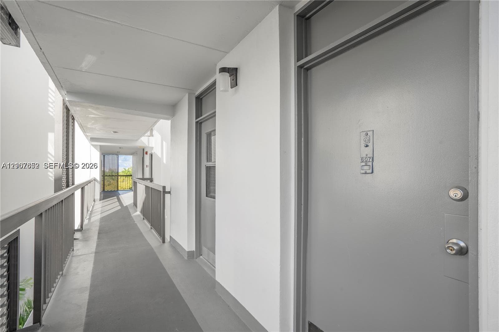 Covered exterior walkway featuring a solid-panel door, wall-mounted light fixture, and metal railing