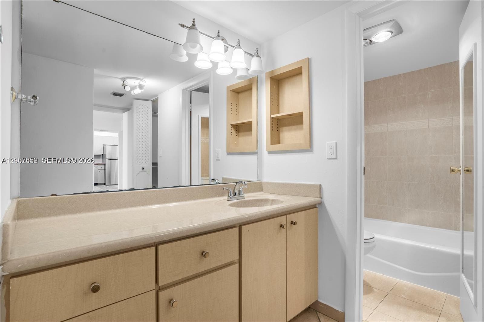 Bathroom featuring a vanity with integrated countertop and sink, light wood-finish cabinetry, built-in wall shelving, a full-width mirror, and a tiled shower-tub enclosure