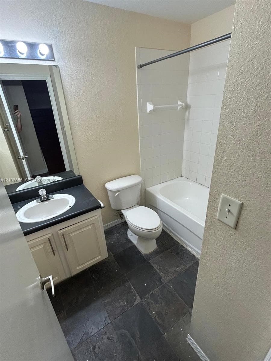 Bathroom featuring dark stone-style tile flooring, vanity with a dark countertop and integrated sink, full-width mirror, and a tub-shower combination with white wall tile