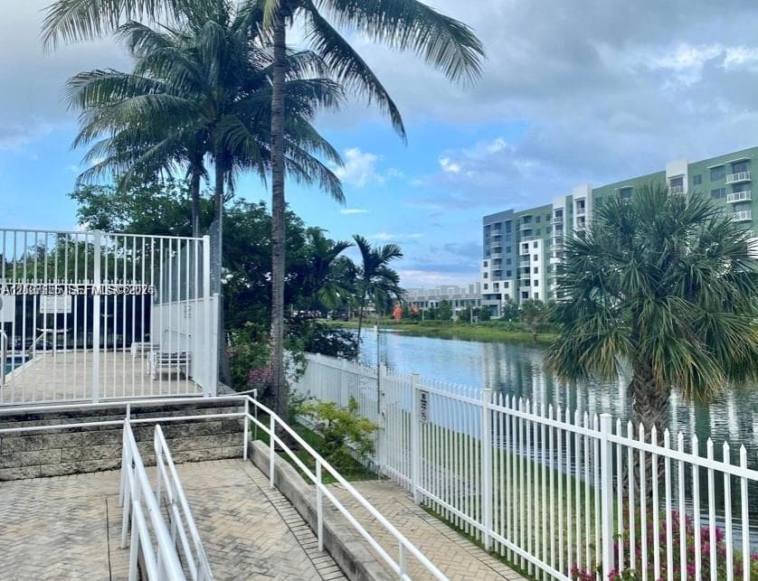 Waterfront outdoor area featuring concrete paver decking, white metal fencing, mature palm trees, and a tiered retaining wall