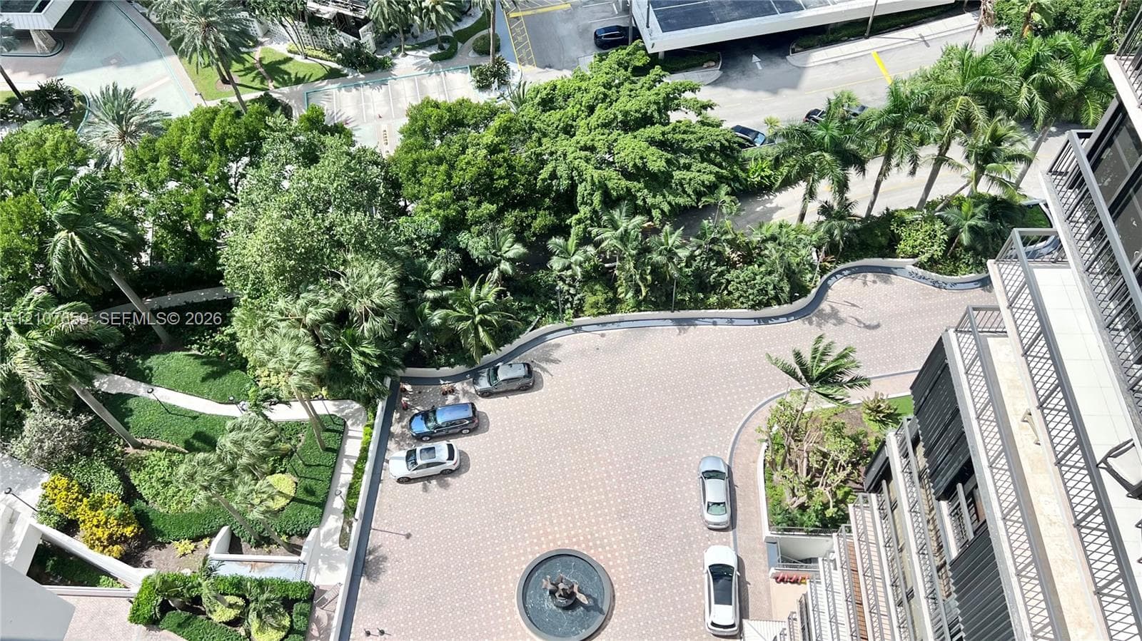 Elevated perspective of the property grounds featuring a paved circular motor court, decorative central fountain, manicured perimeter landscaping, and mature palm trees