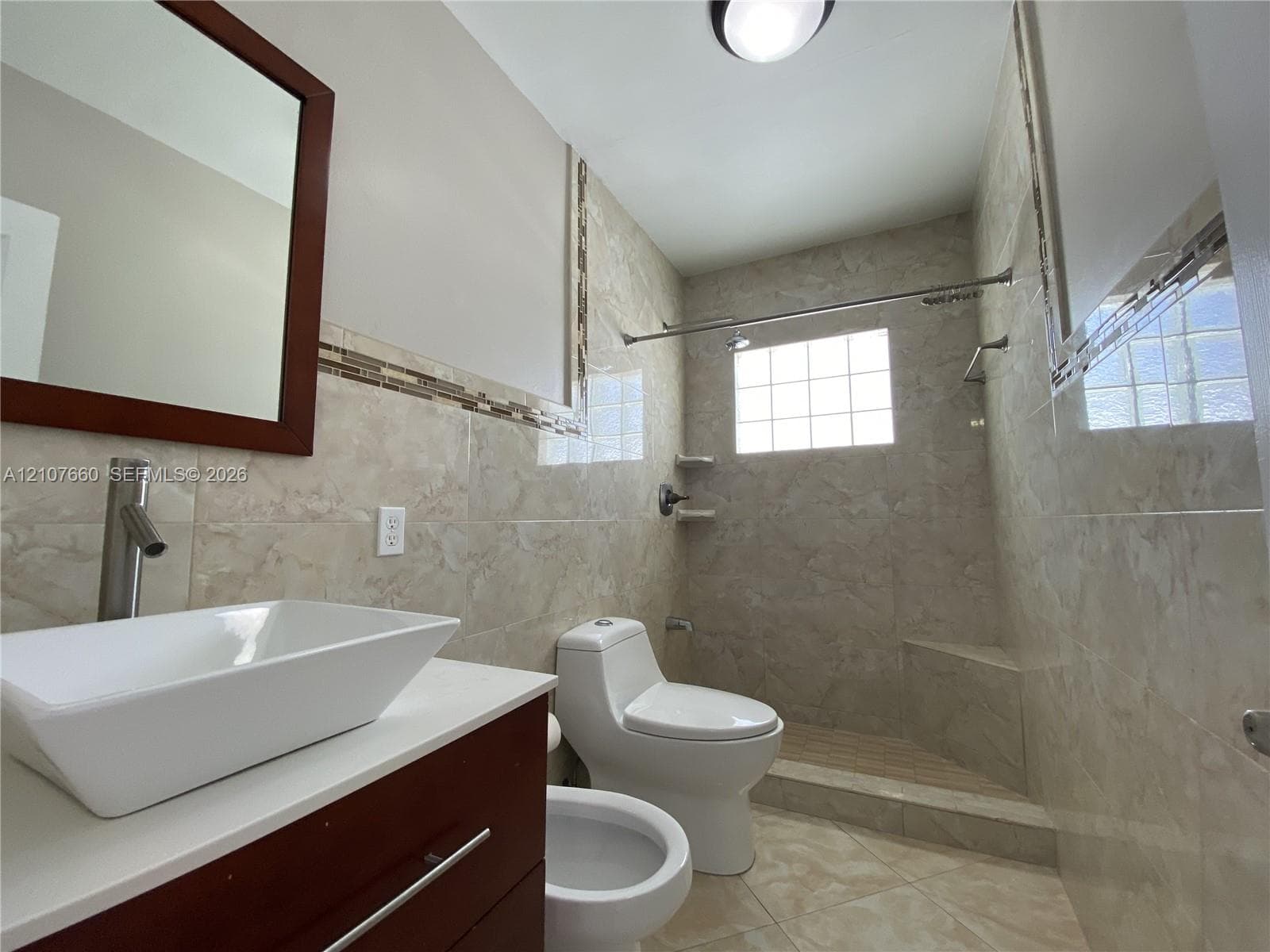 Full-wall stone tiling, rectangular vessel sink on a dark wood-finish vanity, glass block window, and walk-in shower with a built-in corner bench