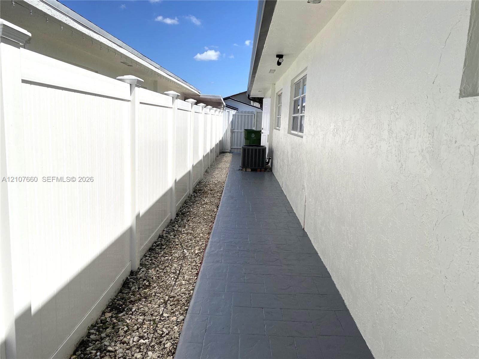 Side yard exterior featuring a white vinyl privacy fence, stamped concrete walkway, and light-toned stucco wall cladding