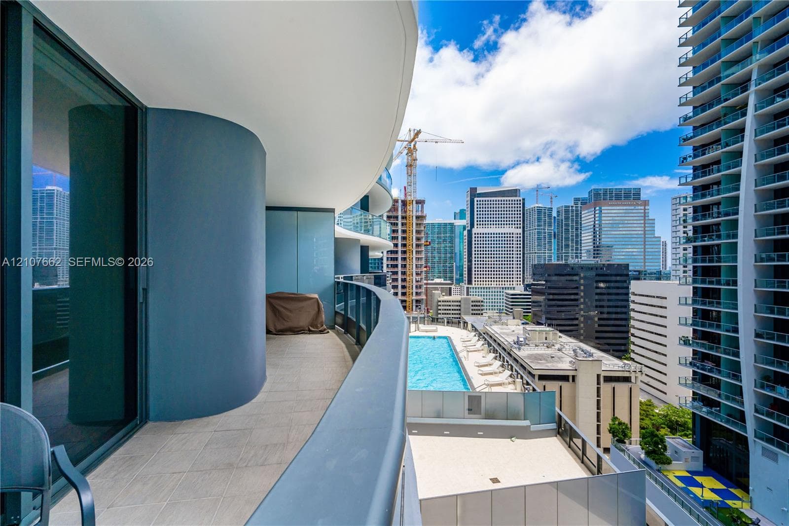 Curved balcony featuring light-toned tile flooring, floor-to-ceiling glass sliding doors, and a glass-paneled railing overlooking an outdoor swimming pool and urban landscape