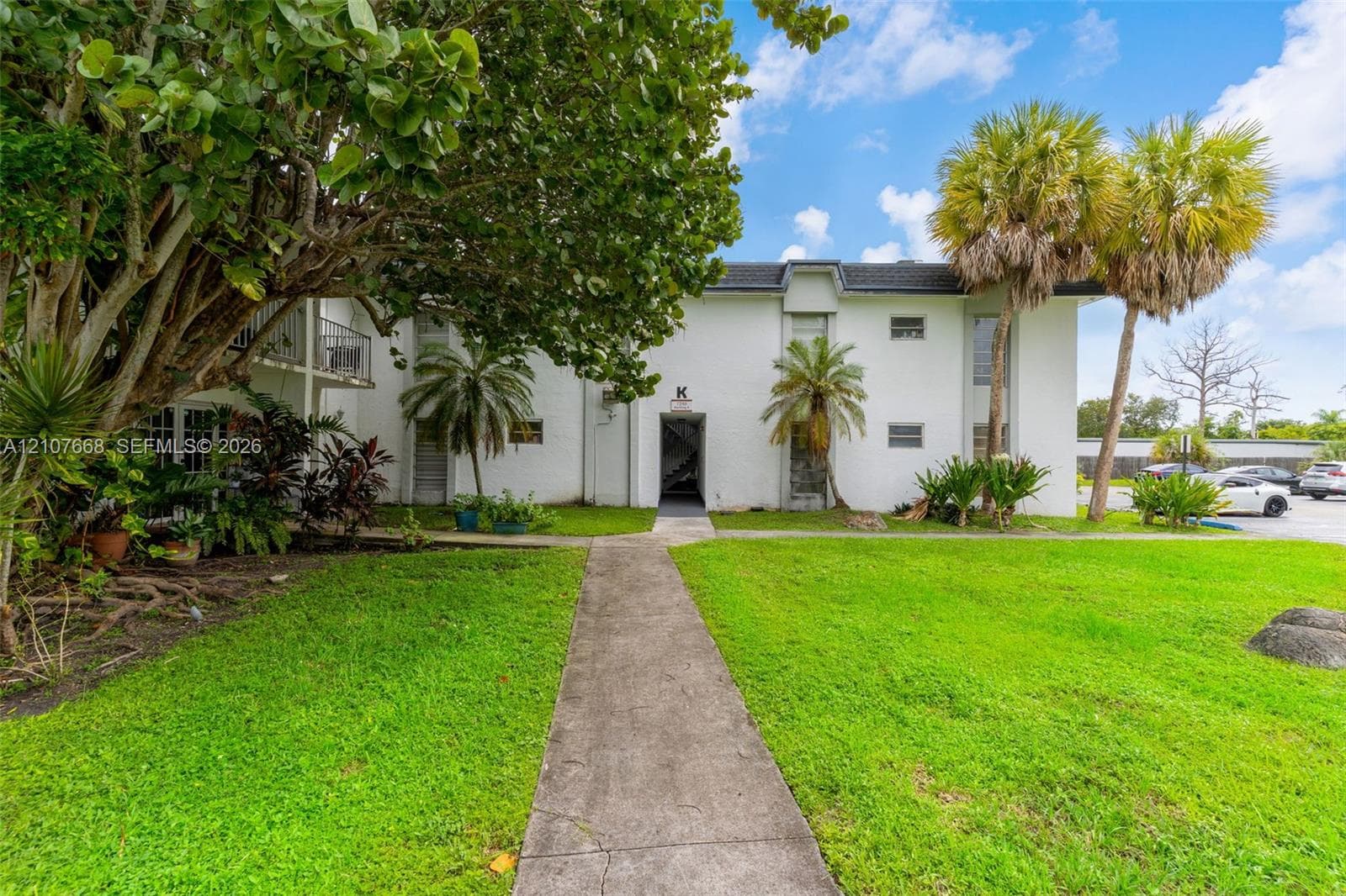 Multi-story exterior featuring white stucco siding, a concrete pathway, mature trees, and palm landscaping