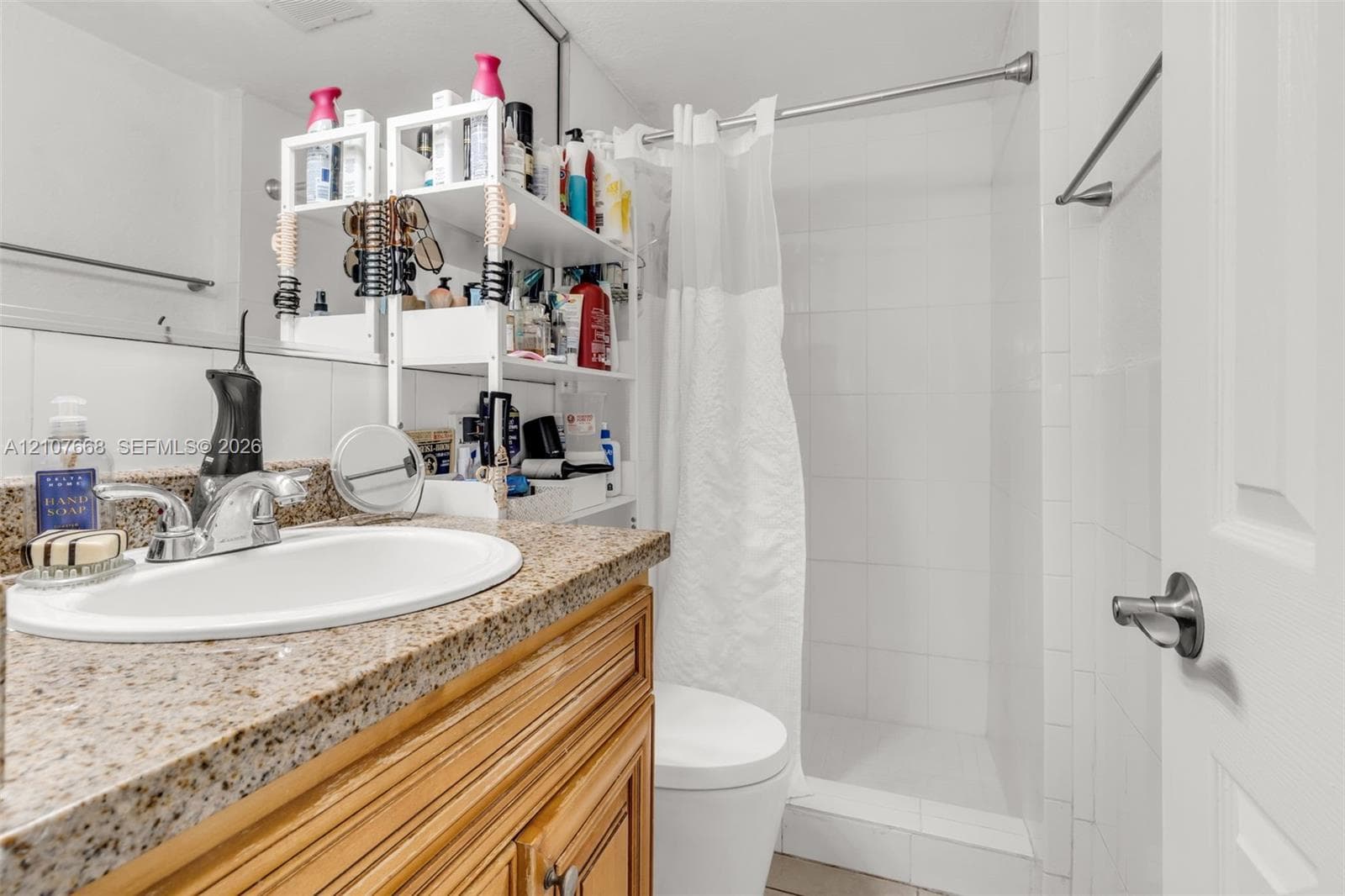 Bathroom featuring a natural stone-finish countertop, undermount sink with chrome-finish faucet, wood cabinetry, and a walk-in shower with white tile surround