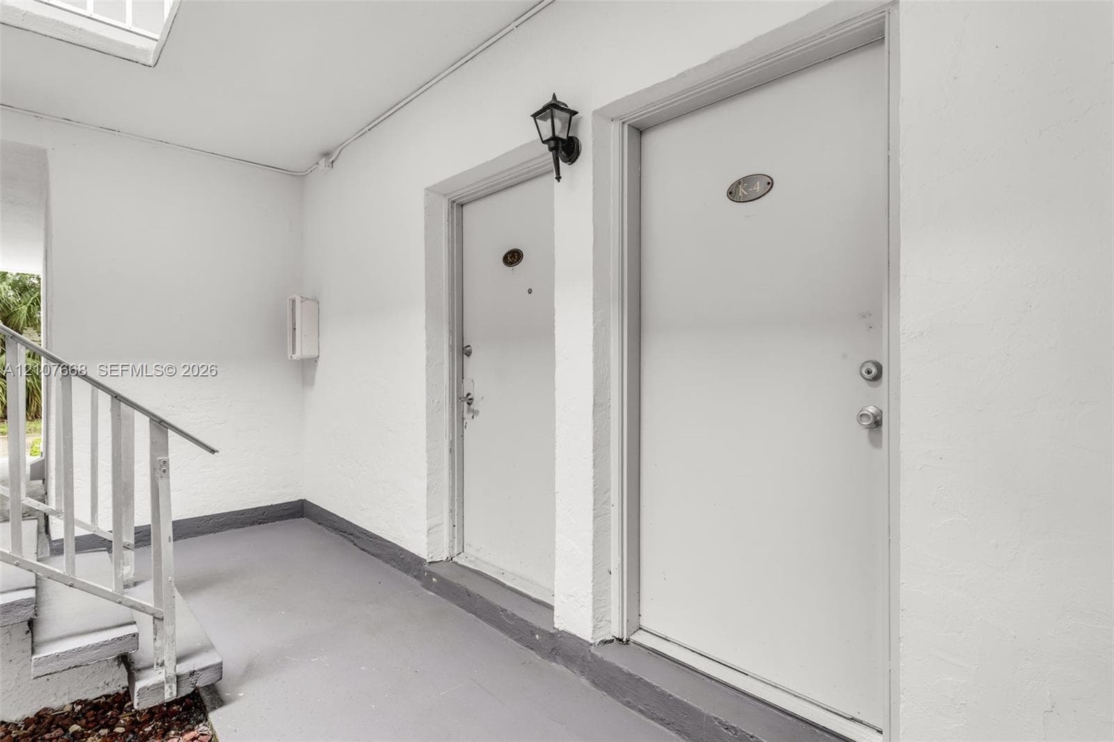 Covered entryway featuring white exterior walls, painted concrete flooring, a metal staircase with railing, and wall-mounted exterior lighting
