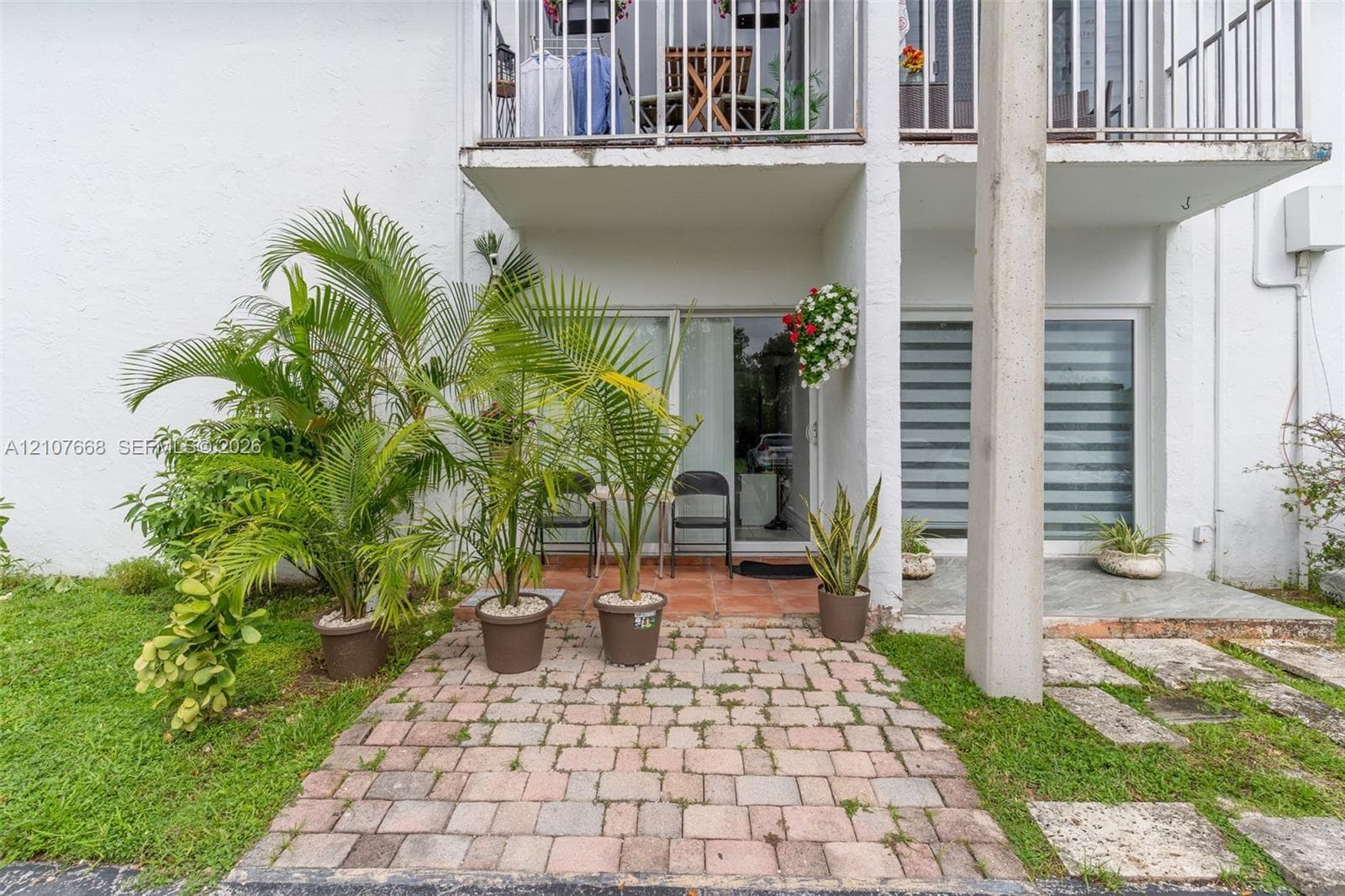 Ground-level patio featuring interlocking concrete pavers, sliding glass entry doors, and a second-story balcony with metal railing