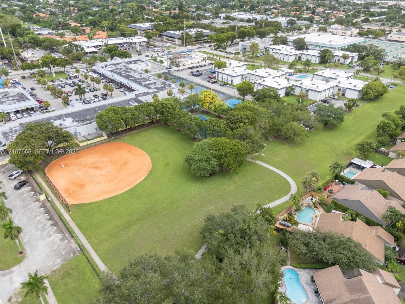 Aerial view of a sprawling recreational area featuring an expansive grass field, a dedicated baseball diamond, paved walkways, and mature trees