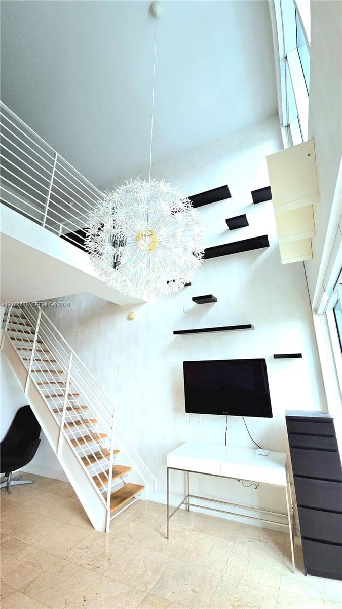 Double-height ceiling featuring a spherical chandelier, white metal staircase with wooden treads, white horizontal railing, and wall-mounted black shelving