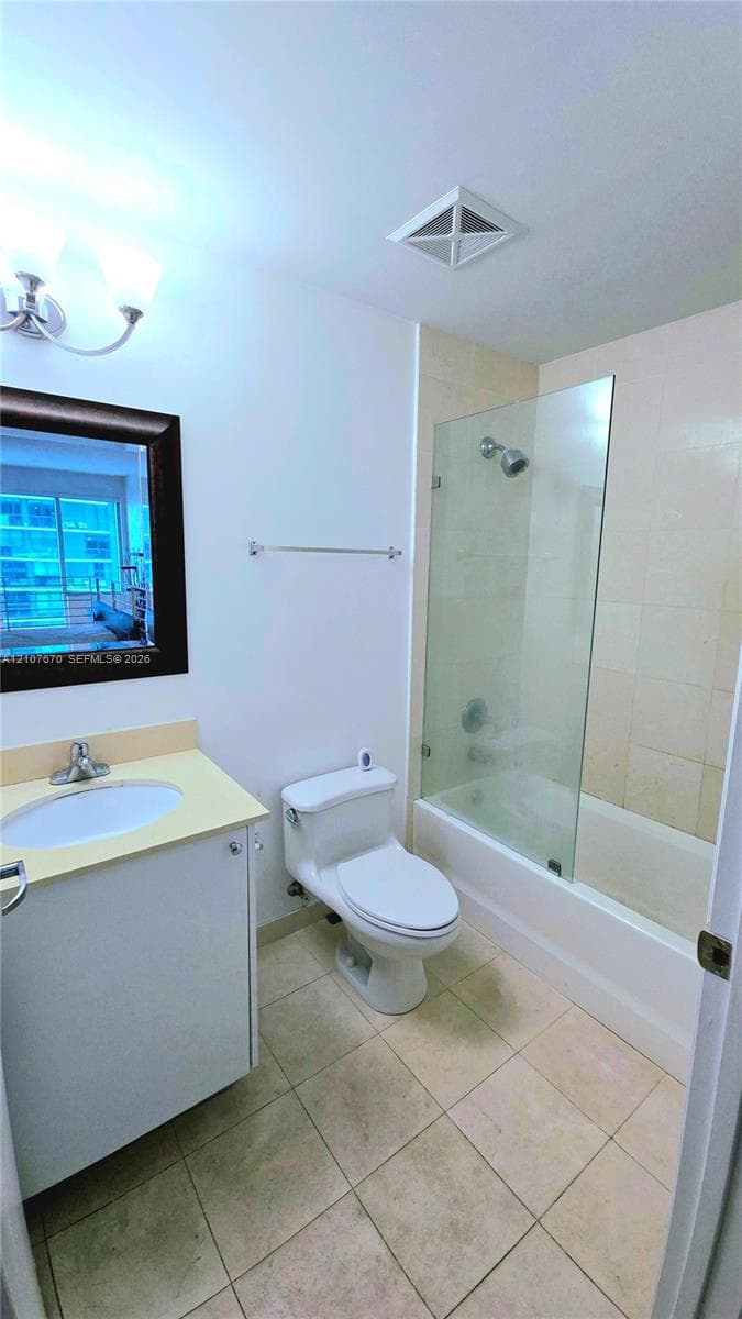 Bathroom featuring ceramic tile flooring, a glass-enclosed shower-tub combination, a vanity with a drop-in basin, and wall-mounted lighting