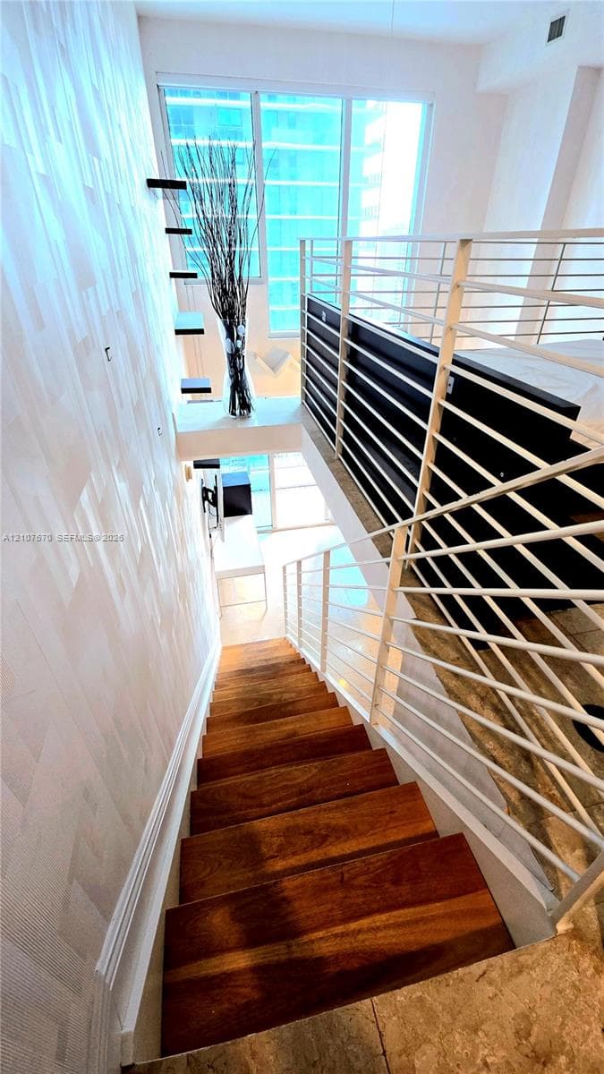 Staircase with wood-finish treads, floor-to-ceiling windows, white horizontal metal railing, and textured wall finish