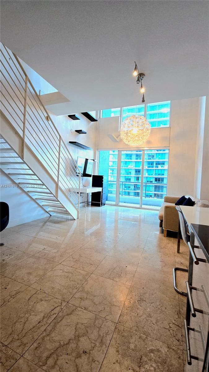 Open-concept living area featuring stone tile flooring, double-height ceilings, floor-to-ceiling windows, a glass-panel balcony door, and a floating metal staircase with integrated railing